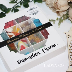 Puede incluir: Una caja de regalo blanca con una ventana transparente, llena de caramelos de colores surtidos. La caja est&aacute; decorada con una cinta negra que dice "Ramadan Kareem" en letras doradas. La caja tambi&eacute;n tiene las palabras "Ramadan Kareem" impresas en la parte delantera.