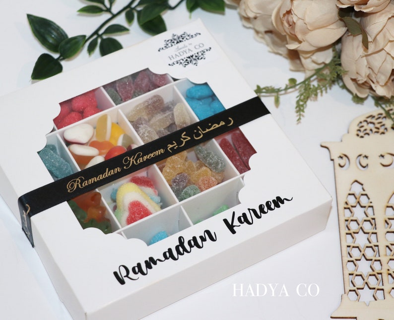 Puede incluir: Una caja de regalo blanca con una ventana transparente, llena de caramelos de colores surtidos. La caja est&aacute; decorada con una cinta negra que dice "Ramadan Kareem" en letras doradas. La caja tambi&eacute;n tiene las palabras "Ramadan Kareem" impresas en la parte delantera.