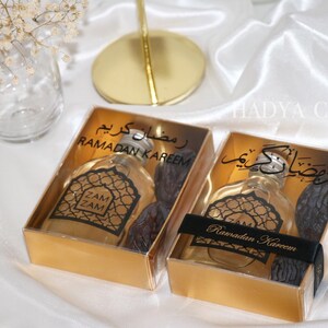 May include: Two gold gift boxes with clear lids containing small bottles of perfume and dates. The boxes are decorated with black text that says "Ramadan Kareem" in Arabic and English.