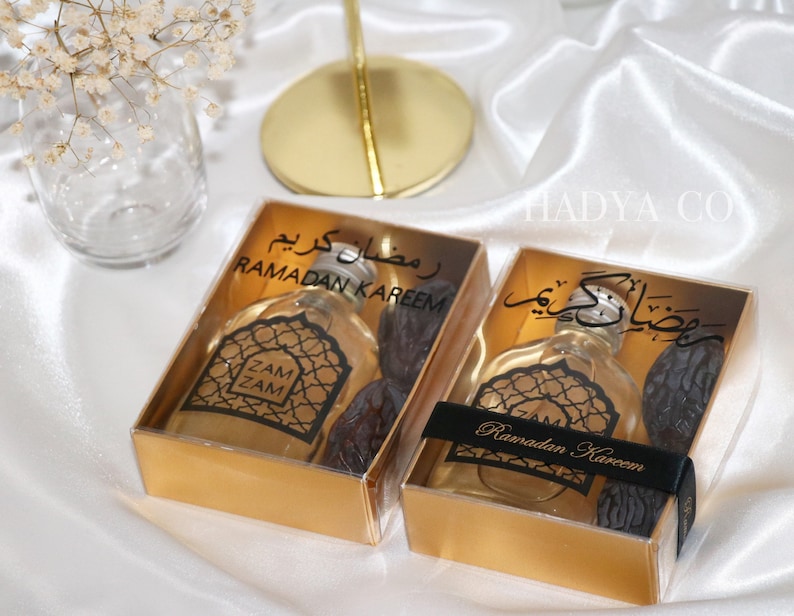 May include: Two gold gift boxes with clear lids containing small bottles of perfume and dates. The boxes are decorated with black text that says "Ramadan Kareem" in Arabic and English.