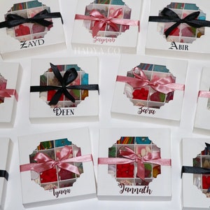 Puede incluir: Cajas de regalo blancas con una ventana transparente que revela caramelos coloridos. Cada caja est&aacute; atada con una cinta de sat&eacute;n negra o rosa y personalizada con un nombre en escritura elegante. Las cajas est&aacute;n dispuestas sobre una superficie blanca.