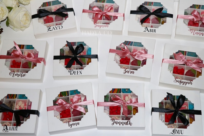 Puede incluir: Cajas de regalo blancas con una ventana transparente que revela caramelos coloridos. Cada caja est&aacute; atada con una cinta de sat&eacute;n negra o rosa y personalizada con un nombre en escritura elegante. Las cajas est&aacute;n dispuestas sobre una superficie blanca.
