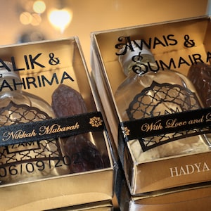 May include: Two gold gift boxes with clear lids, each containing a small bottle of perfume and a date. The boxes are decorated with black ribbon and gold lettering. The text on the boxes reads "Salik & Tahrima Nikkah Mubarak 06/09/2020" and "Awais & Sumaira With Love and Dua's HADYA CO".