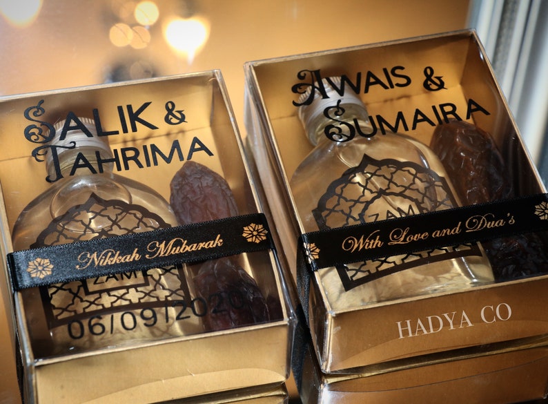May include: Two gold gift boxes with clear lids, each containing a small bottle of perfume and a date. The boxes are decorated with black ribbon and gold lettering. The text on the boxes reads "Salik & Tahrima Nikkah Mubarak 06/09/2020" and "Awais & Sumaira With Love and Dua's HADYA CO".
