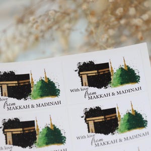 May include: A sheet of white stickers with watercolour illustrations of the Kaaba and a green dome. Each sticker has the text "With love from MAKKAH & MADINAH" in a handwritten font. These stickers are likely for gifts or souvenirs.