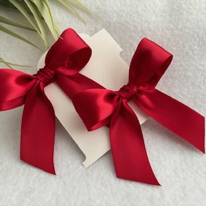 May include: Two vibrant red satin bows, each tied in the center, are displayed on a white background. The bows have long, flowing tails and are attached to small, off-white tags. The image suggests a theme of gift wrapping or crafting.