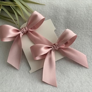May include: Two pink satin bows on a white card. The bows are tied with a knot in the center and have long tails.
