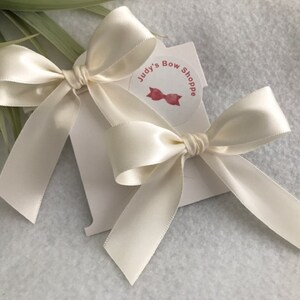May include: Two ivory satin bows tied with a simple knot. The bows are on a white card with the text "Judy's Bow Shoppe" and a red bow illustration.
