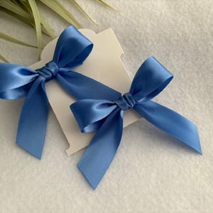 May include: Two blue satin ribbon bows on a white card. The bows are tied in a simple knot and have long tails.