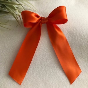 Könnte beinhalten: Eine große orangefarbene Schleife aus Satin mit langen Bändern. Die Schleife ist mit einem klassischen Knoten gebunden und die Bänder fallen herab.
