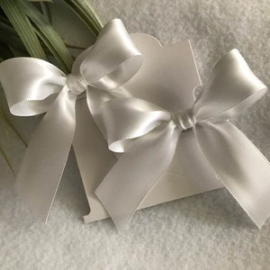 May include: Two white satin ribbon bows attached to a white card. The bows are tied in a simple knot and have long tails.