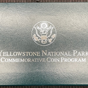 May include: A dark teal box with the text "Yellowstone National Park Commemorative Coin Program" in silver lettering. The box features a silver emblem and a small sticker with the number "1999".