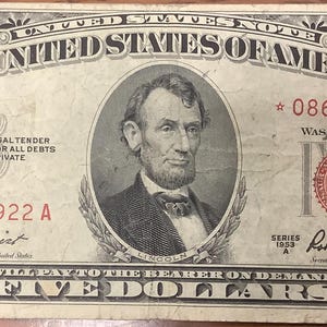 Può includere: Una banconota vintage da cinque dollari degli Stati Uniti. La banconota presenta un ritratto di Abraham Lincoln in una cornice ovale. Il testo "THE UNITED STATES OF AMERICA" è stampato in alto. La banconota ha il numero di serie 08671922 A ed è datata 1953.