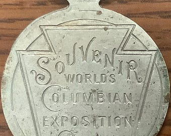 May include: Silver-toned metal souvenir token with a loop at the top. The token features the text "SOUVENIR WORLD'S COLUMBIAN EXPOSITION CHICAGO 1893".