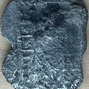 May include: A silver-coloured metal coin with an irregular shape and a worn surface. The coin has a raised design on one side, but the details are difficult to make out.