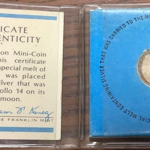 Puede incluir: Una mini-moneda de edición limitada en un estuche protector, acompañada de un certificado de autenticidad. El certificado es blanco con detalles azules y texto. La moneda es plateada y fue transportada a bordo del Apolo 14. El estuche azul dice: "Contiene plata que fue transportada a la luna en el vuelo del Apolo 14."