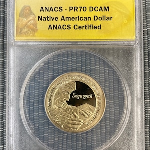 May include: A gold-colored coin in a clear plastic holder. The coin features a portrait of Sequoyah, a Native American who created the Cherokee syllabary, and the text "United States of America" and "Sequoyah". The holder has a yellow label with the text "2017-S ANACS - PR70 DCAM Native American Dollar ANACS Certified $1".