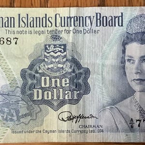 May include: A one-dollar bill from the Cayman Islands, featuring a portrait of a woman wearing a tiara and jewelry. The bill has the text "Cayman Islands Currency Board" and "One Dollar". The serial number is 774687. The bill has blue and green accents.