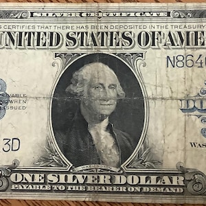 May include: A vintage one-dollar silver certificate from the United States. The bill features a portrait of George Washington and the text "ONE SILVER DOLLAR." The bill is aged, with visible wear and tear.