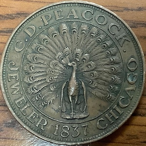 May include: A bronze-colored coin featuring a peacock with its tail feathers fully displayed. The coin is inscribed with "C.D. PEACOCK JEWELER CHICAGO 1837" around the perimeter. The peacock is the central design element.