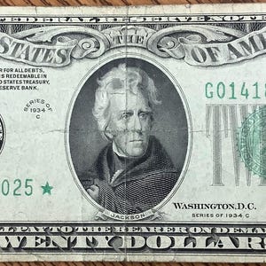 Pode incluir: Uma nota de vinte dólares dos Estados Unidos, com um retrato de Andrew Jackson. A nota é principalmente verde e preta, com o texto "Twenty Dollars" em destaque. O número de série é G01418025.