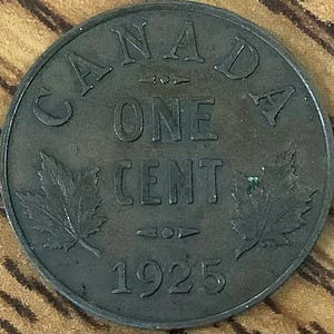 Può includere: Una moneta canadese da un centesimo, scura e circolare, del 1925. La moneta presenta la scritta "CANADA" in alto, "ONE CENT" al centro e l'anno "1925" in basso. Due foglie d'acero affiancano le parole. La moneta ha un aspetto usurato e invecchiato.