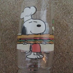 May include: Clear glass featuring Snoopy as a chef holding a large sandwich. Snoopy is wearing a white chef's hat and red shorts. The sandwich has layers of bread, lettuce, and meat. The glass has a wide base and a straight, cylindrical shape.