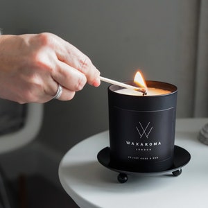 May include: A black candle with a white label that reads "Waxaroma London Velvet Rose & Oud" is being lit with a match. The candle is sitting on a black metal base with three legs.