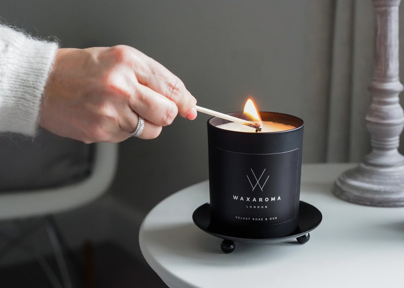 May include: A black candle with a white label that reads "Waxaroma London Velvet Rose & Oud" is being lit with a match. The candle is sitting on a black metal base with three legs.