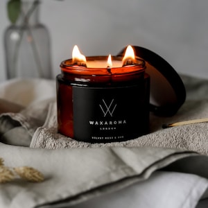 May include: A brown glass candle with three wicks burning. The candle has a black label with the text "WAXAROMA LONDON" and "VELVET ROSE & OUD".
