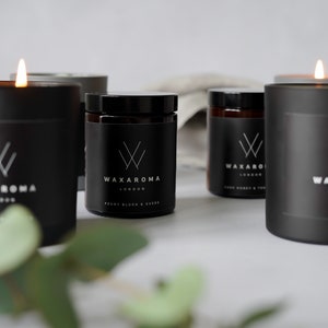 May include: Five black candles with white labels and the text "Waxaroma London" on them. The candles are arranged on a white surface with a green plant in the foreground.