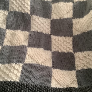 Puede incluir: Una manta de punto con un patrón de tablero de ajedrez en tonos grises y blancos. Los cuadrados tienen diferentes texturas, algunos con un diseño en relieve. La manta tiene un borde gris oscuro.