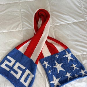 May include: A knitted scarf featuring a red, white, and blue color scheme. One end displays the white text 250 on a blue background, while the other end features white stars on a blue background. The center section has vertical red and white stripes.