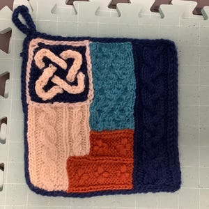 Puede incluir: Salvamanteles cuadrado tejido a mano con un lazo para colgar. Presenta un diseño de nudo celta en crema, punto de cable y secciones texturizadas en melocotón, verde azulado y naranja. El borde y una sección son de color azul marino. Un accesorio de cocina único, hecho a mano.
