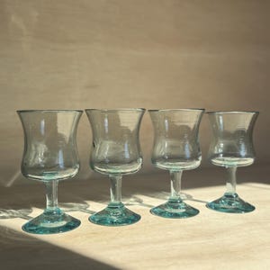 May include: Four clear glass goblets with teal-colored stems and bases are arranged on a light-colored wooden surface. The goblets have a flared bowl shape and a classic stem design. The lighting creates shadows, highlighting the glass's transparency.