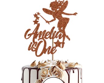 May include: A copper glitter fairy cake topper with the words "Amelia is One" and a star. The topper is on a white cake with chocolate frosting and meringue.