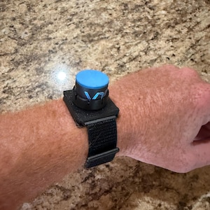 Könnte beinhalten: Ein schwarzes Armband mit einer blauen kreisförmigen Komponente und den Buchstaben "V1" in Blau. Das Armband wird mit einem Klettverschluss befestigt. Das Gerät wird am Handgelenk getragen.