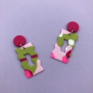 Op de afbeelding: Een paar abstracte oorbellen met roze en groene patronen en rode accenten. De oorbellen zijn gemaakt van polymer clay en hebben een uniek, modern ontwerp.