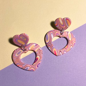 Op de afbeelding: Een paar hartvormige oorbellen met een swirl-patroon in tinten roze, paars en geel. De oorbellen zijn gemaakt van polymer clay en hebben een zilveren haak.