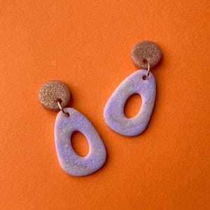 Op de afbeelding: Een paar handgemaakte oorbellen met een lavendel- en wit gemarmerd ontwerp. Elke oorbel heeft een druppelvorm met een cirkelvormige uitsparing en een kleine, ronde, glinsterende stud. De oorbellen zijn opgehangen aan goudkleurige haken.