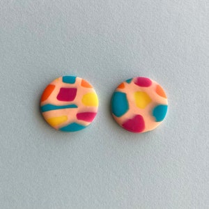 Op de afbeelding: Een paar ronde, roze oorbellen met een kleurrijk abstract patroon. Het patroon bevat geel, blauw, rood en oranje.