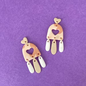 Op de afbeelding: Een paar roze en witte hartvormige oorhangers met gouden accenten. De oorhangers hebben een hartvormige uitsparing en zijn versierd met kleine witte en gouden stippen.