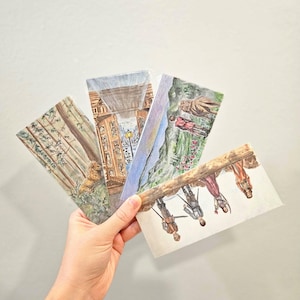 May include: A hand holds a fan of watercolor postcards. Each card features a different scene: a forest, a building with columns, a mountain landscape, and figures on a log. The cards have a light, airy feel.