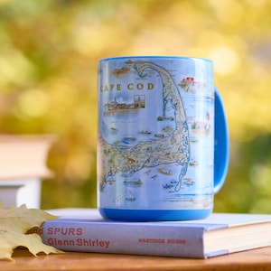 May include: A blue ceramic mug displaying a map of Cape Cod, Massachusetts, with detailed illustrations of landmarks. The mug has a blue handle and base. The mug is resting on a book titled "And Spurs" by Glenn Shirley.