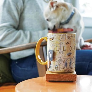 May include: A yellow mug with a map of Yellowstone National Park printed on it. The mug is sitting on a wooden coaster. The text "YELLOW" is visible on the mug.