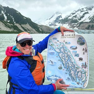 May include: A rectangular poster titled Alaska's Inside Passage featuring a detailed illustrated map of the region, including drawings of wildlife, glaciers, and totem poles, held up against a backdrop of snowy mountains and glacial water.