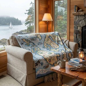 May include: A light blue throw blanket featuring a map of the San Juan Islands, draped over a beige sofa. The blanket has illustrations of marine life and mountains. A mug and books are on a wooden table.