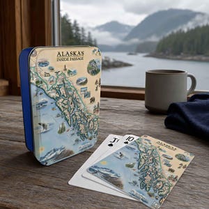 May include: A rectangular blue metal tin featuring a map titled ALASKA'S INSIDE PASSAGE with illustrations of wildlife and landscapes, accompanied by a deck of playing cards displaying the same map design on the top card face.