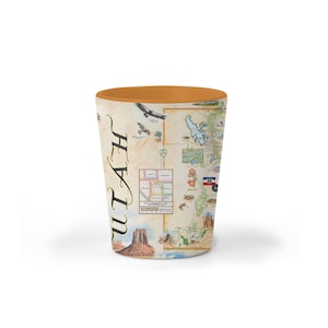 May include: A white ceramic shot glass with a map of Utah and the word "Utah" printed on the side. The map features various landmarks and national parks, including Arches National Park, Zion National Park, and Canyonlands National Park. The shot glass has a brown rim.