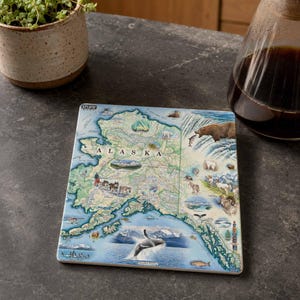 Puede incluir: Un posavasos cuadrado que muestra un mapa detallado de Alaska con el texto ALASKA impreso en el centro. El mapa está decorado con ilustraciones de un oso pardo, una ballena, montañas, glaciares y fauna diversa sobre un fondo azul.